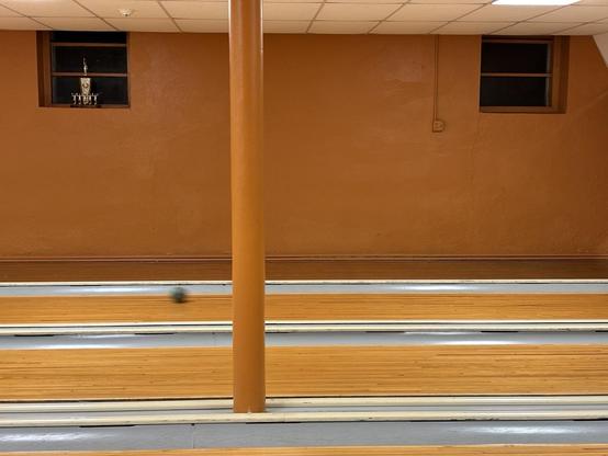 View across 3 bowling lanes with a ball rolling down the far lane.  Ochre walls and pole.  2 windows, one with a trophy on the sill.