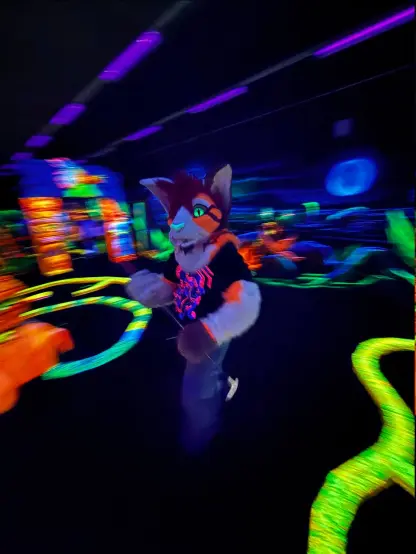A cat partial fursuiter ion a blacklit indoor minigolf course. Not only are the surroundings glowing under the UV light but also the fursuit itself has a bit of an orange glow!