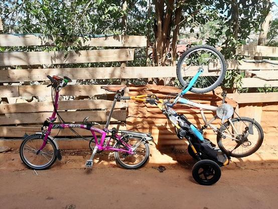A bike towing a bike on a bike trailer. A Brompton bike, pulling a Burley Travoy trailer with a child's bike strapped to it, upside down with wheels in the air.
