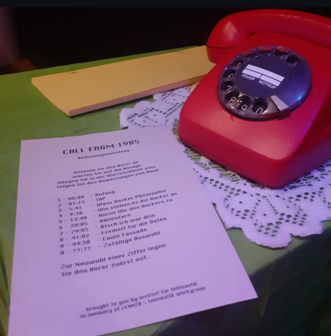 Ein grüner Tisch, auf ihm ein rotes Wahlscheibentelefon auf einer Rüschendecke und daneben eine Bedienungsanleitung