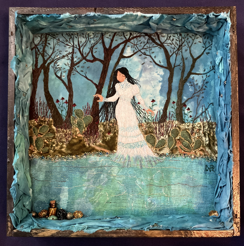 A dark wooden box containing an embroidered panel of a faceless woman with long black hair in a white gown, drifting through the edge of a river in front of a bank growing with cacti, wildflowers, and mesquite trees.