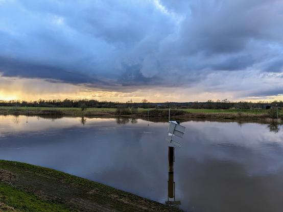Standing on the bank of a river, a bent waterway traffic sign is in the foreground. Just above the trees on the horizon a golden sunset is seen. Above it, a dark cloudy orchestra, some actively downpouring.
