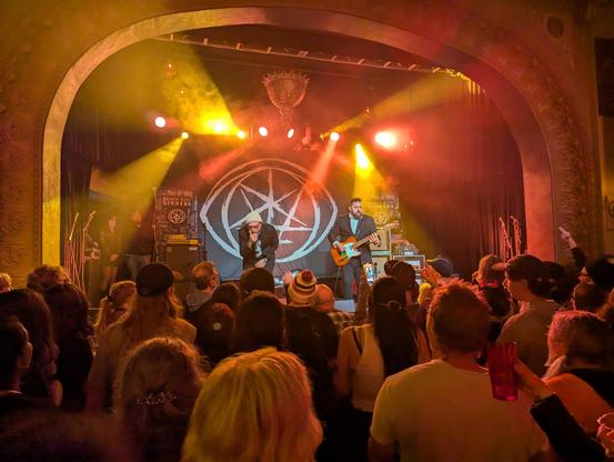 A photo of a three piece band, consisting of a vocalist, guitarist, and DJ
