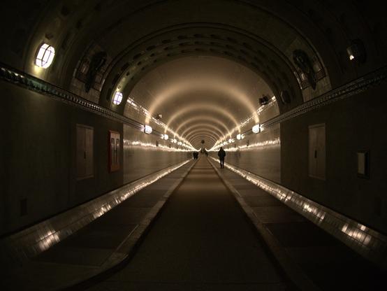Am Beginn des alten Elbtunnels wurde in die Röhre reinfotografiert. Sie ist gleichmäßig beleuchtet und bietet ein harmonisches, symmetrisches Motiv. In der Ferne sind dunkel Fußgänger zu erkennen.