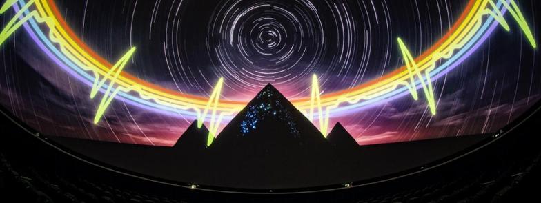 Publicity image for the NSC Creatve show of Pink Floyd’s The Dark Side Of The Moon. Features three dark pyramids silhouetted against a purple sky. Behind the pyramids is a band of the six colours of the spectrum of visible light, and the green band of colour zig zags at intervals, as in a visualisation of a heart beat. In the sky, short curved lines of light make up circles in the sky, as in time lapse photos of stars at night, which streak across the sky as the earth turns.