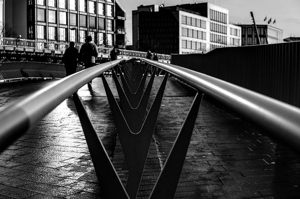 A black and white photo of a bridge railing in the foreground, composed of long, sleek, metallic handrails and dark, V-shaped metal supports. Two people walk in the background, slightly blurred, on a wet, brick paved walkway. Modern buildings are visible in the far background. Thank you @altbot@fuzzies.wtf