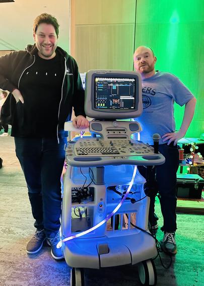 Zwei Männer stehen neben einem medizinischen Ultraschallgerät. Ein Mann trägt einen schwarzen Hoodie und lächelt, während der andere ein graues Hemd trägt. Die Maschine ist mit einem Bildschirm ausgestattet, der Daten anzeigt, und hat eine bunte Lichtdekoration um sie herum.