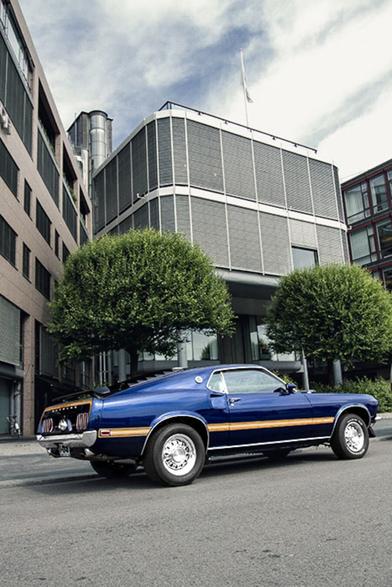 This image features a vintage blue Ford Mustang, parked in front of a grey building with white trim, surrounded by lush greenery. The car's sleek design is accentuated by its dark blue paint job, while the surrounding trees add a touch of natural beauty to the scene.