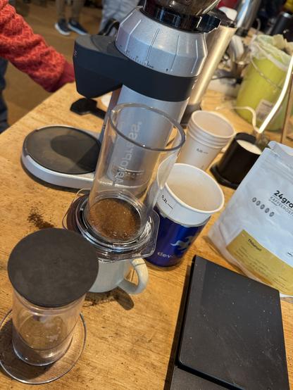 Eine Kaffeezubereitung mit einer Silbermühle, einem Glasbrühgefäß mit gemahlenem Kaffee, einem Keramikbecher, einem Pappbecher und einer Tüte Kaffeebohnen auf einem Holztisch. Eine digitale Waage ist ebenfalls sichtbar.