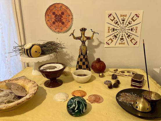 A pagan altar with a Minoan Snake Goddess figurine, a white offering stand with a crocheted bee an dried lavender on it, an incense bowl, a pomegranate, a large yellow bowl full of bay leaves and linen thread, three seashells, a green handblown glass orb, and several smaller objects. They all sit on a yellow altar cloth. On the wall behind them are two Minoan-themed mandala style designs drawn in colored ink.