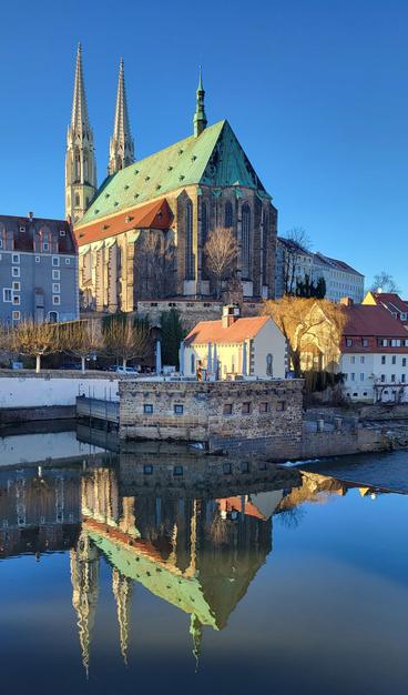 Das Bild zeigt eine malerische Ansicht einer Kirche mit zwei hohen Türmen und einem grün-kupferfarbenen Dach. Die Kirche steht am Ufer eines ruhigen Flusses, der ihr Spiegelbild perfekt widerspiegelt.  Um die Kirche herum gruppieren sich weitere Gebäude, darunter niedrige Häuser mit roten Dächern. Der Himmel ist klar und blau, die Sonne scheint, was für eine ruhige und friedliche Atmosphäre sorgt. Das Gesamtbild vermittelt einen Eindruck von Geschichte und Architektur in einer europäischen Stadt.
―