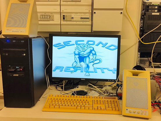 Messy PC setup. Black medium tower case with CD-ROM and Gotek/CF as well as 3.5" floppy drive. 24" Dell LCD monitor. Old yellow keyboard. Magnificent Trust SoundWave speakers. Second Reality demo by Future Crew on screen.