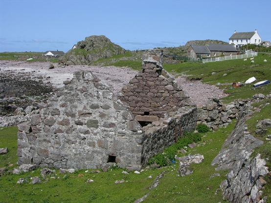 The image shows a ruined stone cottage in the bottom left of the frame with a beach beyond it.The beach faces to the left but the tide is out.  There are some boats pulled up above the beach and there are further buildings, not ruined, on the horizon in the middle distance including a white house on the skyline in the upper right of the frame. The sky is blue.