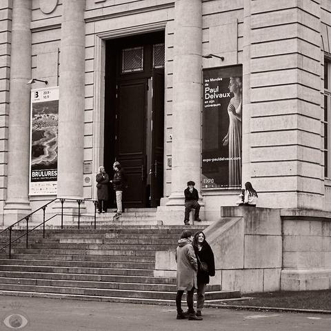 The entrance of La Boverie, the museum where the exhibition is held. Three pillars are seen above a stone stairwell. On each side of the entrance is a poster.People are standing and sitting in front.