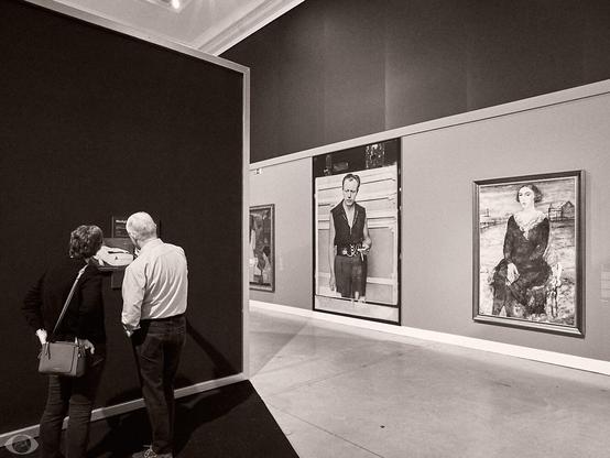A woman and a man standing in front of a painting of Modigliani, only partly visible. At the wall to the right 2 big paintings of Delvaux, and, in the middle, a big photo of the master.