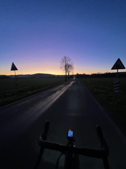 A silhouette of a cyclist's handlebars and bike computer is visible in the foreground, with an empty road extending into the distance. On either side of the road, there are road signs and two trees against a colorful dawn sky transitioning from deep blue to shades