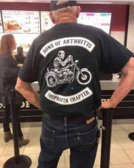 Man at a take out line, from behind, black ball cap, thinning  hair, hands on sore hips, black T-shirt and blue jeans. His shirt shows a motorcycle driving skeleton with a helmot . It reads, "SONS OF ARTHRITIS - IBUPROFEN CHAPTER"