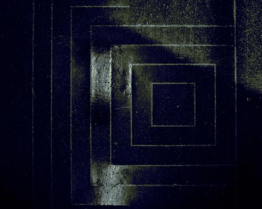 The image shows a section of a black floor tile with a reflective surface. The tile features a pattern of nested, squared shapes, each fitting within the other.