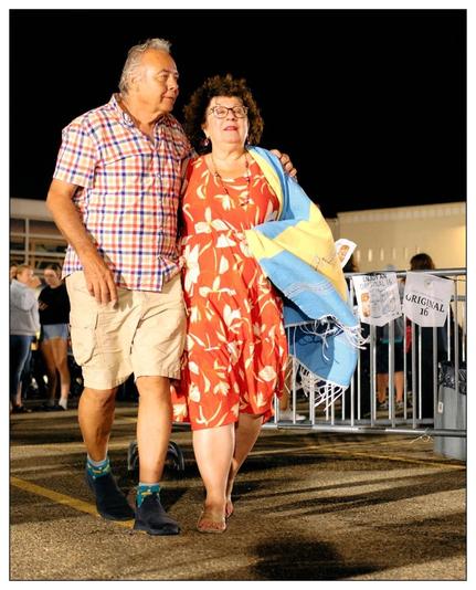 The image shows a middle-aged couple walking closely together at what appears to be an evening outdoor event or gathering. The man is wearing a plaid short-sleeve shirt, khaki shorts, and dark blue shoes, with his arm around the woman. The woman sports a red and white floral dress, glasses, and is carrying a large blue beach towel along with a smaller item, possibly a jacket or a shirt. Both individuals are smiling and seem to be enjoying their time. In the background, there are other people milling about, and there is also a fence with advertisements hanging, suggesting a commercial or public setting, possibly near a beach or during a festival.