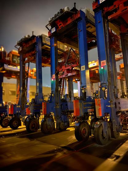 Abgebildet sind drei Portalhubwagen. Es sind Flurförderfahrzeuge zum Umschlag von ISO-Containern. Sie werden vornehmlich als Transportfahrzeug auf Containerterminals in Häfen eingesetzt. Aus einem blauen Metall Rahmengestell und einer dazwischen mittig aufgehängten Hubvorrichtung (Spreader), die mit Hubwinden vertikal bewegt werden kann. Das blaue Rahmengestell ist mit einem Fahrwerk mit jeweils vorne und hinten vier schwarzen Rädern ausgestattet. Die Führerkabine ist oben hinten rechts des Rahmens angeflanscht in orange mit Glasöffnungen. Das Bild wurde bei Nacht aufgenommen.