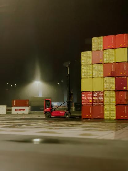Abgebildet ist eine Nachtszene. Vorne im Bild befindet sich ein grauer asphaltierter Boden. Mittig im Bild befindet sich ein rotes Fahrzeug zum bewegen der Container. Das Fahrzeug steht seitlich nach rechts. Die schwarze Hubvorrichtung wurde nach oben geklappt. Rechts davon Athene viele rote, orange und gelbe auf und nebeneinander gestapelte Metall Container. Über dem Fahrzeug leuchtet eine Laterne die Nachtszene aus. Der Himmel ist dunkel bewölkt. Leichter Nebel umhüllt die Szene im Hintergrund.