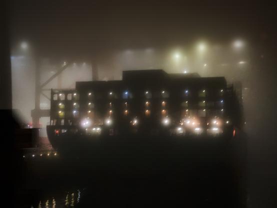 Mittig im Bild steht am Hamburger Hafen ein großes Containerschiff. Die Aufnahme entstand bei Nacht und starkem Nebel. Daher wirkt das Bild etwas verschwommen. Die Lichter auf dem Schiff, die Spiegelung vor dem Schiff auf dem Wasser, sowie die verschwommenen Kreise der Laternen im Hintergrund wirken diffus. Alle ist leicht verschwommen.