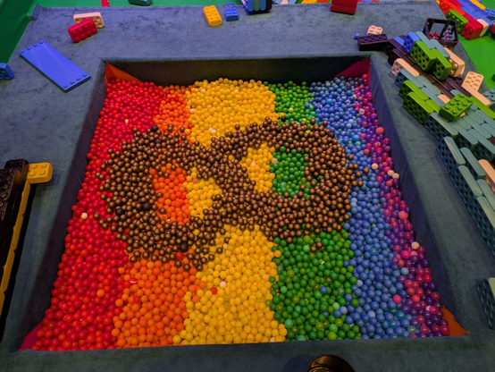 A ballpit sorted by color making a golden infinity sign on top of a rainbow surrounded by huge lego bricks.