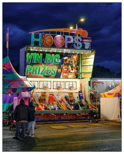 This image shows a vibrant scene from a carnival or fair at dusk, based on the moody sky and artificial lighting. Featured prominently is a "Hoops" game booth where players can shoot basketballs into hoops to win prizes. The booth is adorned with colorful lights and basketball-themed decorations, including images of basketball players and oversized representations of basketballs. The top of the booth displays signs with enticing phrases like "WIN BIG PRIZES" and "ONE IN, YOU WIN". Additionally, there are some plush toys and NBA jerseys displayed as potential prizes for winners.

Several individuals are visible in the image. In the foreground, a group of people is standing and observing the game, and in the background, a game attendant can be seen sitting on a chair, likely managing or supervising the booth. The atmosphere suggests a fun and energetic evening setting typical of such events.
