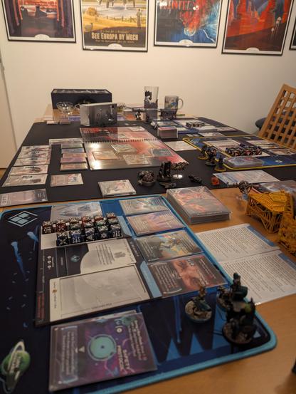 Table full over boardgame stuff. Dice, playmats, minis, cards. Some chocolates in the background, and a mug and glass.