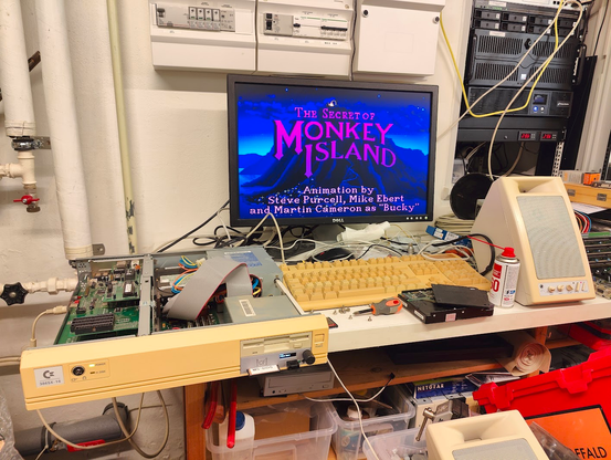 My lab. Open Commodore 386-SX16 dekstop computer visible. Gotek and CF combo device in one of the 3.5" bays. LCD monitor connected, showing the intro from Monkey Island. Glorious Trust speakers connected.