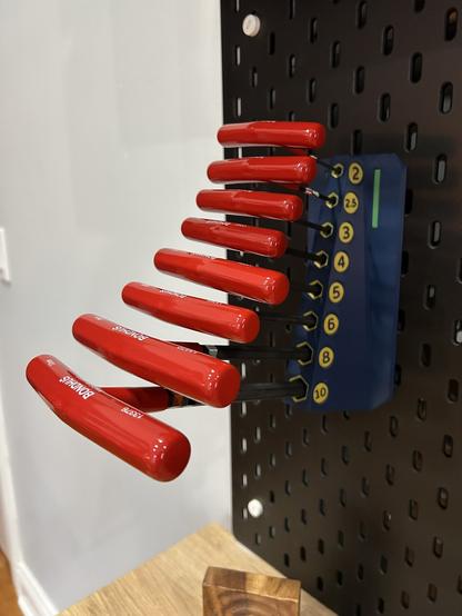 A tall blue mount is hanging on a black IKEA SKÅDIS pegboard.  It holds 8 T-handle hex wrenches at tilted up at a 15 degree angle from horizontal.  Along the side of the mount, the wrench sizes in millimeters are printed in gold, from 2 at the top through 10 at the bottom.  Each wrench is inserted into a hexagonal hole, and the holes are outlined in gold to make them easier to find.  Near the top, one of the hooks is visible as a thin green rectangle.  The wrenches have red soft plastic handles, and the brand, Bondhuis, is printed on them.