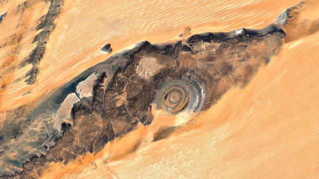 A satellite picture of a desertic landscape. In the center is a large, dark, almost spiral-shaped geological formation that contrasts sharply with the lighter-colored sand dunes surrounding it. All around, the sand dunes stretch out, exhibiting a variety of textures and shades. The overall color palette is warm, with sandy browns and oranges dominating, offset by the darker hues of the central rock formation.