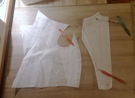 A top down photo of two paper pattern pieces on a desk, poorly lit. An array of pens is littered on top of it, to hold it down. All of the front and the side back are in one continuous and very shapely piece, the back being quite narrow with one singular princess seam running along its edge. Most of the arm's eye is on the front and side piece, giving a fun little scooped out hole in it. That's where an arm goes! It's generally not as clear in pattern pieces. A large floppy prototype lapel shape is dangling off the centre front and at the very least it's massive. Two darts at the side front of waist, also.
