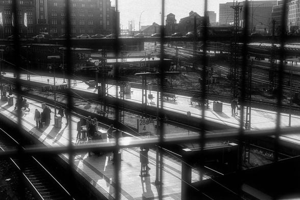 Here's a detailed alt-text description of the image: A black and white photo shows a view through a metal grid fence of a busy train station platform. Several people are visible on the platform, some standing in small groups, others walking individually. The platform is long and stretches into the distance, with train tracks and more platforms visible beyond. The background includes buildings, cars, and a complex network of train tracks. A sign with the numbers "288 4" is partially visible on one of the platforms. The overall scene is one of urban activity, viewed from a slightly elevated perspective. The strong sunlight creates distinct shadows on the platform. Provided by @altbot@fuzzies.wtf