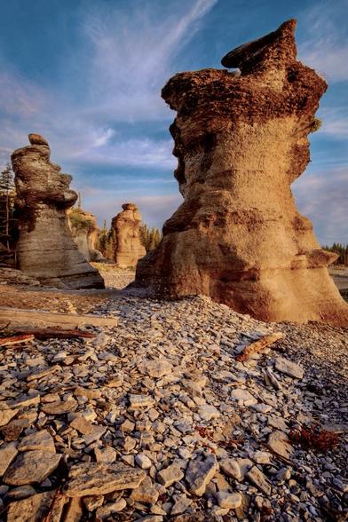 Vividly colorful vertical shot of one large seastack at right, and two smaller seastacks at farther left, bathed in warm rising sunlight on a rocky beach underneath a bright blue partly cloudy sky.