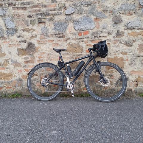 My bike, a black gravel bike with fenders, a handlebar bag and 2 thermos flasks, leaning against a grey-brown stone wall.