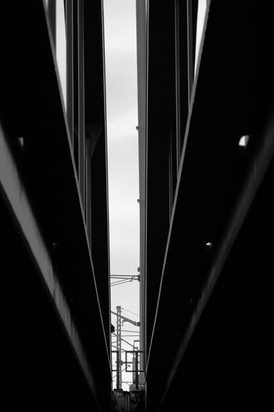 A black and white photograph shows a narrow view between two large, dark metal structures. The structures are made of thick, parallel beams, creating a strong sense of depth. In the distance, through the gap, are visible thinner metal poles and what appears to be some kind of overhead railway signaling equipment. The sky is visible in the background, appearing light and somewhat overcast. Provided by @altbot@fuzzies.wtf