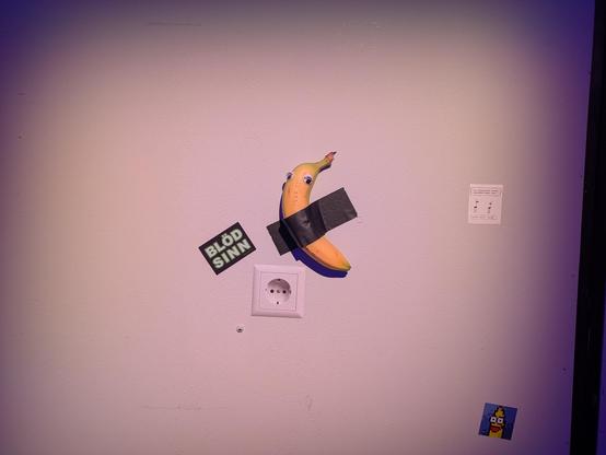 A banana taped to a wall next to a sticker power socket and a sticker which says "blödsinn"
