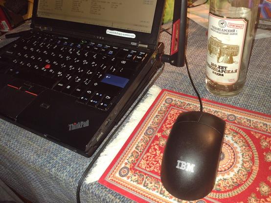 Modded Thinkpad X220 with an old IBM black mouse connected to it. Mouse has two keys and a ball.
Under the mouse there is a mouse pad made as red carpet with white fringes, popular in Slavic world.
There is a bottle of Cheboxary vodka on the background.