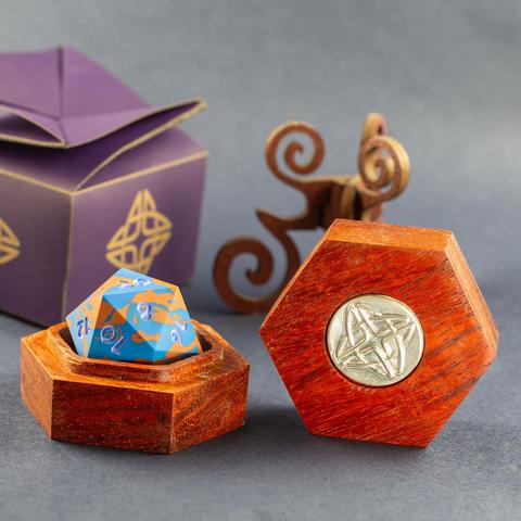 Padauk hardwood hexagonal box with a silver Celtic knotwork medallion in the lid, open with a blue and copper 42mm Jesmonite 20-sided die inside. An ornate spiraled walnut stand and a purple box with the same knotwork motif are visible in the background.