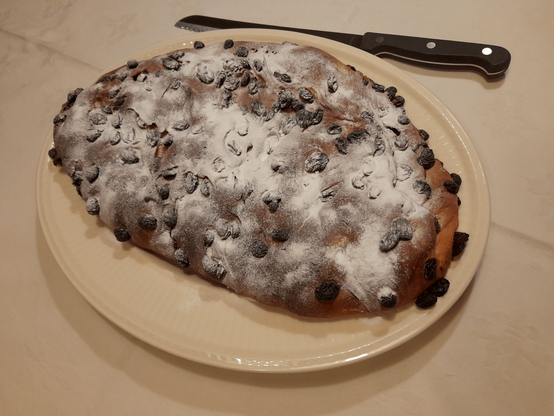 A Dutch Christmas bread on a plate. It is very low, contains raisins and is covered in powdered sugar. Next to the plate is a large knife.