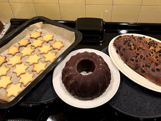 A baking tray filled with star-shaped cookies on top of an oven, as well as a chocolate wreath cake and Dutch christmas bread (without powdered sugar) on plates.