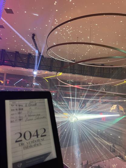 Foto des eBooks von 2042 - Die verbotene Bibliothek auf einem eReader, der vor der Lichtkunst in der Eingangshalle des CCH in bunten Farben leuchtet