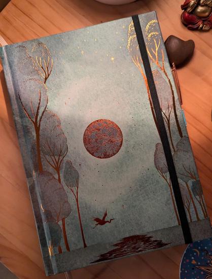 A teal green journal with a bronze full moon, trees, and a river with a heron in flight on the cover.