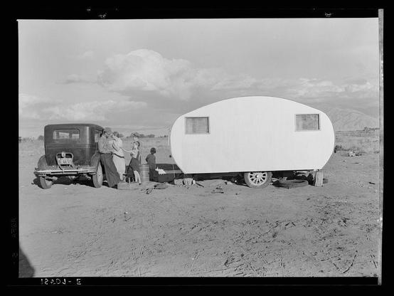 A black and white photograph depicts an older model car parked alongside a trailer in what appears to be a dry, open field. The sky is partly cloudy with hints of mountains visible on the horizon line. Attached to the rear of this vehicle is a small white trailer, which has two windows and seems simplistic in design.

Several individuals are standing near the back door of the car; they appear engaged in some kind of interaction or activity together. One person appears to be reaching out towards another individual who stands behind them. The ground looks dusty with sparse vegetation around.

This image is titled "Migratory family from Louisiana, been in California eighteen months now on Works Progress Administration (WPA), receives fifty-five dollars a month. No house available in nearest town under twenty dollars a month." It suggests that the people depicted are part of a migratory or transient group living with limited resources and facing challenges such as finding housing while supported by government programs like WPA during an uncertain time period.

The photograph likely captures a moment from the lives of migrant workers, reflecting their hardships and perseverance in pursuit of better opportunities.