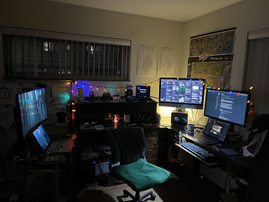 A darkish room with several computer monitors on a large desk. Colored lights across the top..