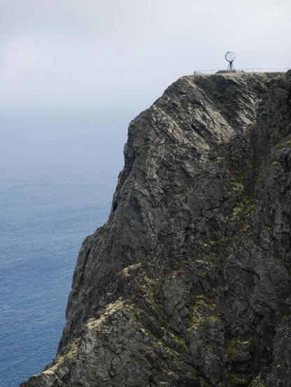 Hohe Steilklippen, links unten ist das Meer zu sehen, rechts oben steht auf den Klippen eine große Skulptur aus Metall die die Weltkugel mit den Längen- und Breitengraden darstellt.