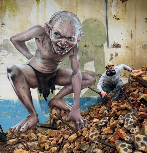 A stunning 3D graffiti artwork by French artist SCAF, featuring Gollum from The Lord of the Rings. The mural is painted on a crumbling wall, showing Gollum crouched on rubble with a lifelike expression of intensity. Cleverly integrated props and painted details enhance the realism, making Gollum appear ready to leap out of the scene. SCAF himself is posed next to the artwork, blending seamlessly into the setting.