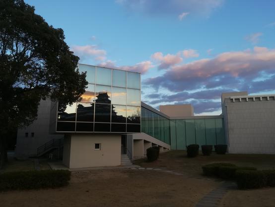 Himeji Castle reflected in a grid of windows at about 4:45pm on December 29, 2024. The sun has set. The reflected sky is turquoise with orange clouds, while the clouds in the sky behind the museum are pink and violet.