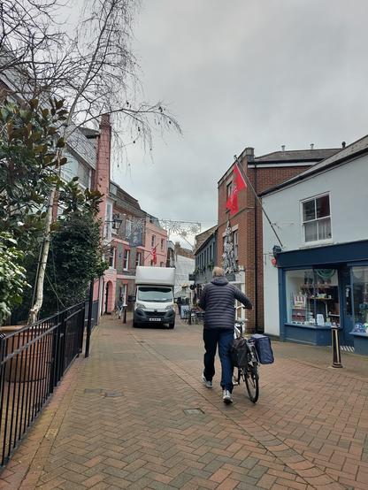 a high top van parked in a pedestrian area (the wrong way for the flow of traffic when it is allowed for access). it squeezes up against pavement tables for a cafe and leaves a narrow gap for pedestrians to walk past. a man walks his bike towards the van